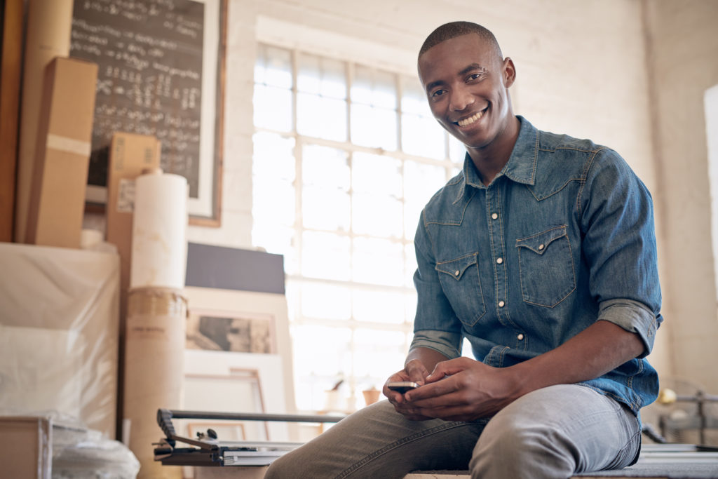 Low angle portrait of a handsome young business owner of African descent, smiling at the camera while holding is mobile phone and sitting on a workbench in his studio workshop
